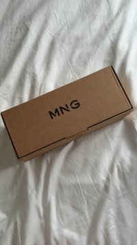 Scatola rettangolare di cartone marrone con la scritta "MNG" stampata in nero sulla parte superiore, poggiata su una superficie di tessuto bianco spiegazzato.