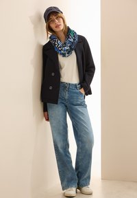 Marineblå dobbeltbrystet jakke, hvid top, lysblå vide jeans, mønstrede blå tørklæde og hvide sneakers, parret med en marineblå kasket.