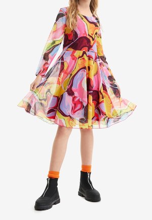 Kobieta ubrana w sukienkę za kolano z długim rękawem o kolorowym, abstrakcyjnym wzorze, pomarańczowe skarpetki i czarne botki na platformie z zamkiem.