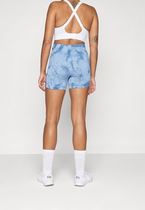 Person der står med ryggen til, iført en hvid sports-bh, blå tie-dye shorts, hvide crew sokker og hvide sneakers mod en ensfarvet baggrund.