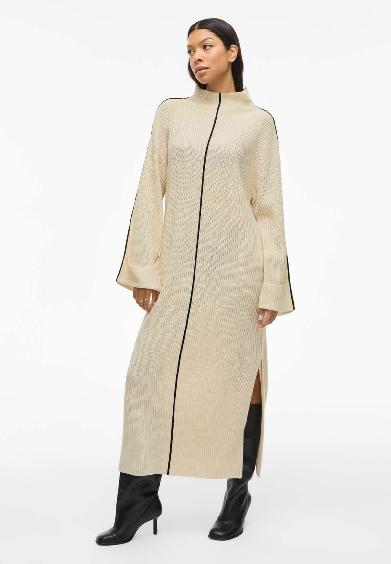 Robe tricot beige avec un col montant, présentant des accents noirs sur les côtés et une fermeture éclair à l'avant. Manches longues, fentes latérales et coupe décontractée.