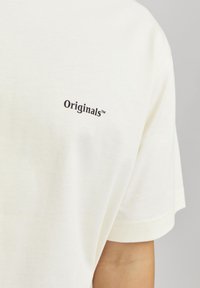 Kremfarget bomulls t-skjorte med korte ermer, med en liten svart "Originals™" logo på brystområdet. Glatt tekstur.