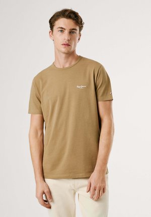Jeune homme à la peau claire et aux cheveux bruns ondulés, portant un t-shirt à manches courtes couleur sable et un pantalon beige clair, debout devant un fond uni.