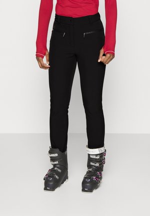 Persoon die zwarte skibroek, zwarte skistappers met zilveren gespen en een rood shirt met lange mouwen draagt, staand op een effen witte vloer.