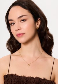 Złoty naszyjnik z okrągłym wisiorkiem, noszony na brązowej, teksturowanej bluzce na cienkich ramiączkach. Włosy ułożone w luźne fale.