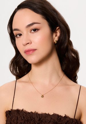 Złoty naszyjnik z okrągłym wisiorkiem, noszony na brązowej, teksturowanej bluzce na cienkich ramiączkach. Włosy ułożone w luźne fale.