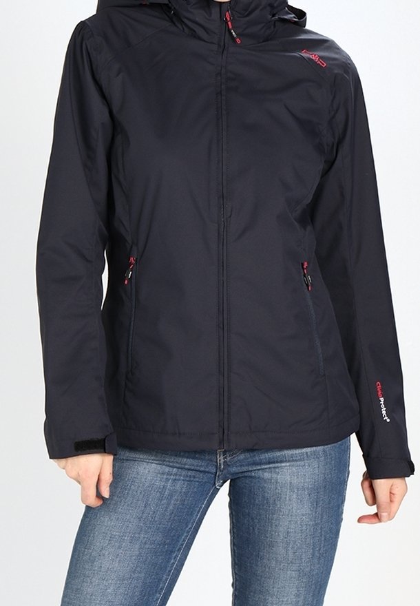 Personne portant une veste à capuche noire zippée avec des tirettes de fermeture éclair rouges et un jean bleu, debout devant un fond uni.