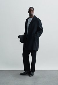 Fekete gyapjú kabát laza siluettel, szürke pulóver és slim fekete nadrág párosítva. Fényes fekete bőr táska a kézben.