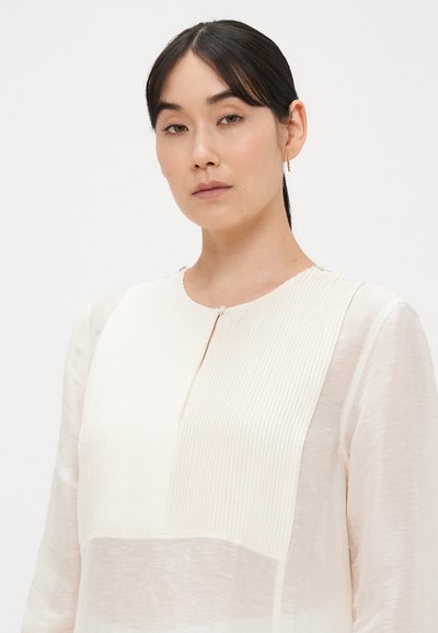 Ελαφριά κρεμ μπλούζα με πιετέ υφή στο επάνω μισό, ελαφριές διαφανείς μανσέτες και στρογγυλή λαιμόκοψη με μικρή λεπτομέρεια κουμπιού.