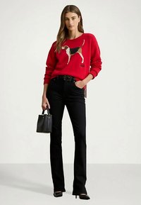 Ženska v rdečem puloverju z motivom beagleja, črnih hlačah z višjim pasom, črnih petah in drži majhno črno torbico.