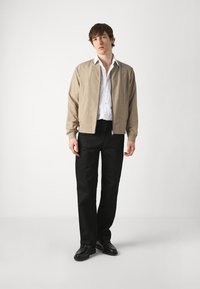 Giubbotto bomber beige con polsini a coste, indossato sopra una camicia bianca con bottoni, abbinato a pantaloni neri a gamba dritta e scarpe nere.