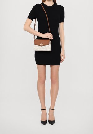Kobieta w czarnej sukience z krótkim rękawem, czarnych szpilkach z paskiem wokół kostki, niosąca brązową i beżową torebkę na ramię z złotym logo jako zapięciem.