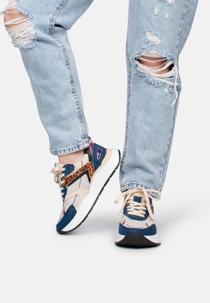 Podarte jasnoniebieskie dżinsy połączone z wielomateriałowymi sneakersami w kolorach beżowym, granatowym, w panterkę oraz z białą podeszwą na neutralnym tle.