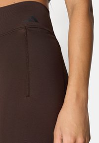 Brune atletikleggings lavet af strækbart stof med flatlock-sømme. Har en høj talje og en sort logoaccent nær toppen.