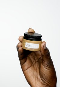 Barattolo di vetro ambrato con coperchio nero contenente balsamo per labbra etichettato "BALSAMO PER LABBRA" e dettagli sugli ingredienti; tenuto in una mano su sfondo bianco.