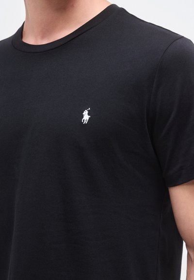 T-shirt de algodão preta com gola redonda, mangas curtas e um pequeno logótipo branco bordado no peito esquerdo. Textura macia.