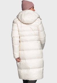 Lange, lichtbeige pufferjas met capuchon, met horizontale gestikte secties en een gladde textuur. Draagt een lichtroze gebreide beanie.