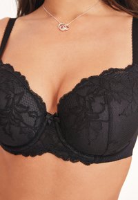 Reggiseno con ferretto in pizzo nero con un design strutturato e a motivi, caratterizzato da un dettaglio a fiocco al centro e bordi smerlati sulle coppe.