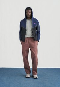 Marineblauw en zwarte fleecejas met rits, gecombineerd met een grijs T-shirt, bordeauxrode joggers en grijze sneakers, alles op een blauwe vloer.