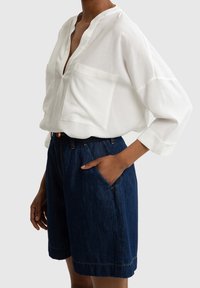 Blusa bianca in lino con scollatura a V e due tasche frontali, abbinata a pantaloni corti in denim scuro con orlo arrotolato e tasche laterali.