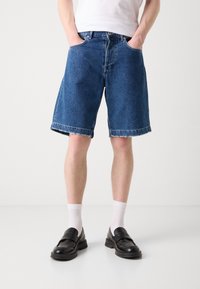 Shorts en denim bleu moyen avec une coupe décontractée, dotés de revers et de poches latérales, portés avec des chaussettes blanches et des mocassins noirs.