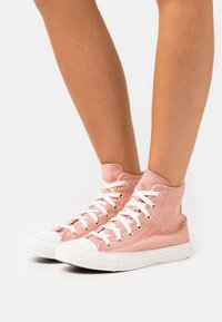 Vaaleanpunaiset korkeavartiset tennarit, joissa on rakenteellinen kangas ja metallinhohtoiset kultapilkkuja, valkoiset nauhat ja kumipohja.