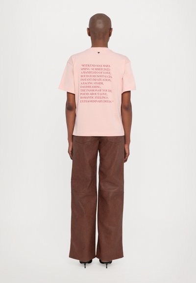 Tricou din bumbac roz deschis cu mâneci scurte și imprimeu text pe spate; asortat cu pantaloni largi maro și pantofi cu toc negri.