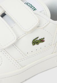 Λευκό δερμάτινο αθλητικό παπούτσι με ιμάντες βέλκρο, με κεντημένο πράσινο λογότυπο κροκοδείλου και διάτρητες λεπτομέρειες στο πλάι.