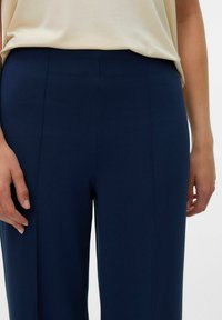 Pantalons sur mesure bleu marine avec une texture lisse, présentant des coutures verticales et une taille haute, associés à un haut beige clair.