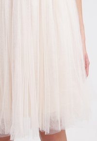 Vaaleanbeige tyllihame kerroksellisella kankaalla, läpikuultavalla tekstuurilla ja A-linjaisella mallilla, jossa on pehmeitä, virtaavia säikeitä.