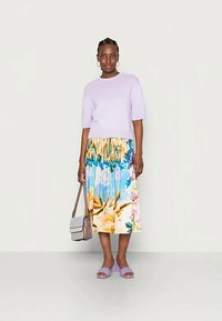 Mulher de pé vestindo um suéter lilás de mangas curtas, uma saia midi floral colorida, sandálias trançadas lilás e carregando uma bolsa bege.