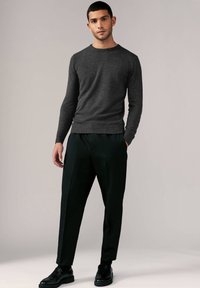 Grauer Strickpullover mit rundem Halsausschnitt, kombiniert mit lockeren schwarzen Hosen und schwarzen Schuhen, vor einem neutralen Hintergrund.