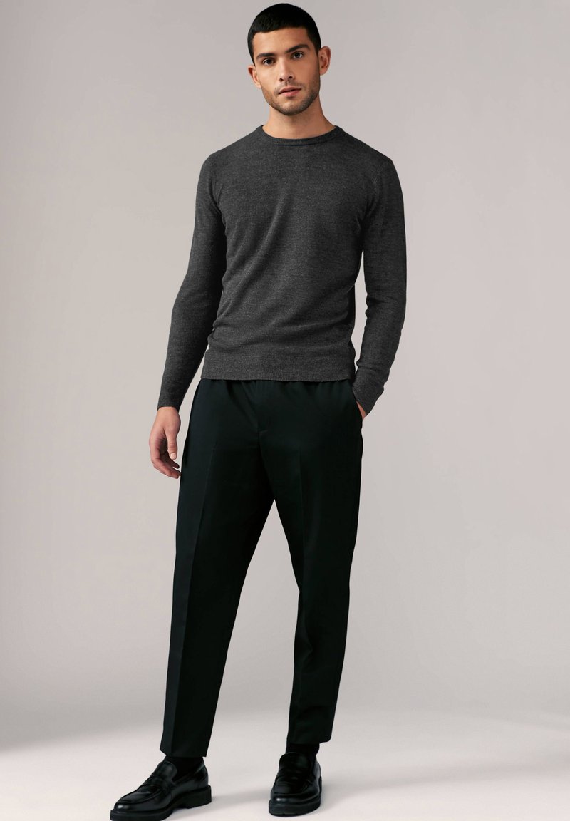 Grauer Strickpullover mit rundem Halsausschnitt, kombiniert mit lockeren schwarzen Hosen und schwarzen Schuhen, vor einem neutralen Hintergrund.
