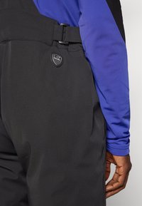 Melnas slēpju bikses ar jostu un logo uzlīmi. Audums ir gluds, un tās ir apvienotas ar zilu garām piedurknēm krekli.