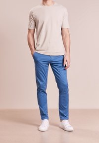 Světlé béžové tričko s krátkým rukávem spárované s modrými slim-fit kalhotami a bílými teniskami, vystavené na neutrálním pozadí.