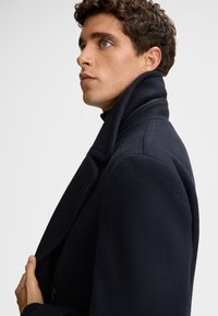 Marine-blauer Wollmantel mit hohem Kragen und strukturierten Schultern. Texturiertes Gewebe und ein einzelner Knopfverschluss sind sichtbar. Seitenansicht.