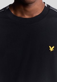 Svart bomull t-shirt med rund hals, har grå accenttråd och en gul broderad örnlogotyp på vänster bröst.