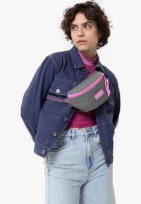 Marsupio grigio con accenti rosa, realizzato in un materiale testurizzato, indossato sopra una giacca blu e una maglietta a pois rosa, dotato di chiusura a zip.