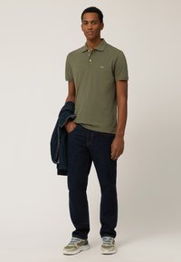 Camisa polo verde oliva con tapeta de tres botones, combinada con jeans azul oscuro. El hombre sostiene una chaqueta azul marino y lleva puesta unas zapatillas grises y blancas.