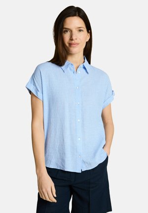 Femme aux cheveux lisses et foncés portant une chemise boutonnée bleu clair à manches courtes et un pantalon foncé, debout avec une main dans la poche.