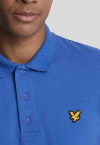 Polo shirt blu in cotone con scollatura a colletto, dotato di tre bottoni e un logo di aquila gialla sul lato sinistro del petto.