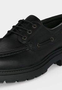 Chaussure bateau en cuir noir avec semelle épaisse à crampons, trois œillets avec lacets noirs et couture moc toe sur un fond clair.
