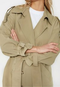 Kobieta z długimi włosami, złożonymi na piersiach, ubrana w beżowy trencz na białą koszulę, kadrowana od podbródka do bioder na jednolitym tle.