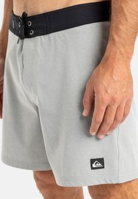 Sivé plavky s čiernym elastickým pásom, so nastaviteľnou šnúrkou a malým logom na ľavej strane.
