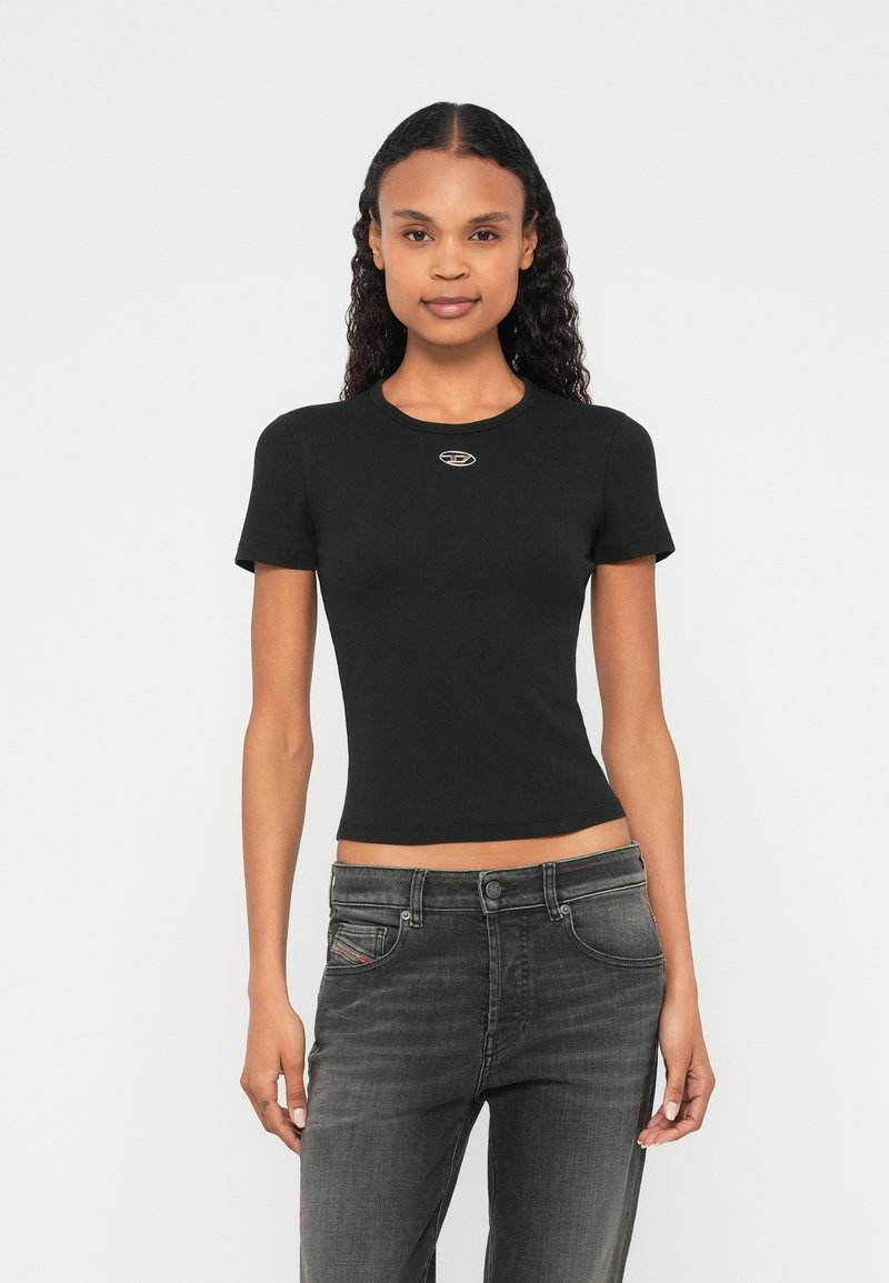 Melnā īsroku cieši piegulošā T-krekls ar apaļu kakla izgriezumu un nelielu sudraba logotipu uz krūtīm; kopā ar pelēkajām džinsiem.