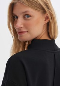 Schwarzes Textilkleidungsstück mit hohem Kragen, das eine glatte Textur und Nahtdetails an Rücken und Schultern aufweist, getragen von einer Person mit hellem Haar.