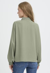 Light green blouse with long sleeves and ruffled collar detail. Soft fabric, slightly loose fit, and buttoned cuffs. Worn with blue jeans.