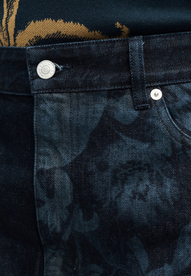 Close-up van donker denim jeans met subtiele bloemmotieven en metalen knopen, gedragen met een zwart-gele top met patroon.