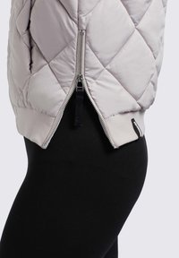 Lichtgrijze doorgestikte jas met een rits aan de zijkant en geribde manchetten, gecombineerd met zwarte leggings. De stof heeft een gladde textuur.