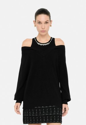 Vrouw in een zwarte off-shoulder trui over een mouwloos topje met een rhinestone halslijn, gecombineerd met een zwart-witte gestructureerde rok.